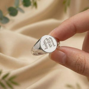 May include: A silver signet ring with a circular face. The face has an engraved letter "M" in a stylish font. The ring is held between fingers against a soft, neutral background.