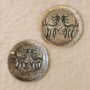 Könnte beinhalten: Zwei Keramikplatten mit gesprenkelter, graubrauner Oberfläche. Jede Platte zeigt eine stilisierte Illustration von zwei Pferden, die sich gegenüberstehen, in Grün und Weiß. Die Teller sind rund und scheinen handgefertigt zu sein.