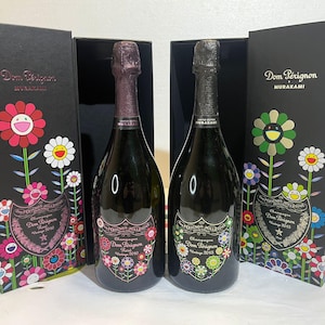Könnte beinhalten: Zwei Flaschen Dom Pérignon Champagner, jeweils in einer schwarzen Schachtel mit farbenfrohen Blumenmustern. Die Flaschen haben Etiketten mit floralen Akzenten und dem Text "Dom Pérignon" und "MURAKAMI". Eine Flasche ist Vintage 2010, die andere Vintage 2015.