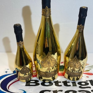 Puede incluir: Tres botellas de champán doradas de diferentes tamaños. Cada botella tiene una tapa negra y una etiqueta dorada con un símbolo de pica. Las botellas están dispuestas sobre una superficie blanca con el texto "Bottlig crea".