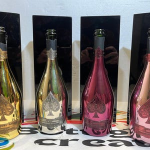 Puede incluir: Cuatro botellas de champán con acabados metálicos: dorado, plateado y dos tonos de rosa. Cada botella tiene una etiqueta negra y un logotipo de pica. Las botellas se exhiben frente a paneles rectangulares negros.