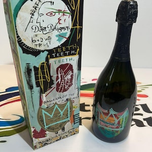 Pu&ograve; includere: Una bottiglia di champagne Dom P&eacute;rignon con una parte superiore in lamina nera e un'etichetta colorata. Accanto, una scatola abbinata con opere d'arte in stile graffiti, tra cui testo e un disegno a corona. La scatola riporta le parole "Dom P&eacute;rignon" e "In God We Trust".