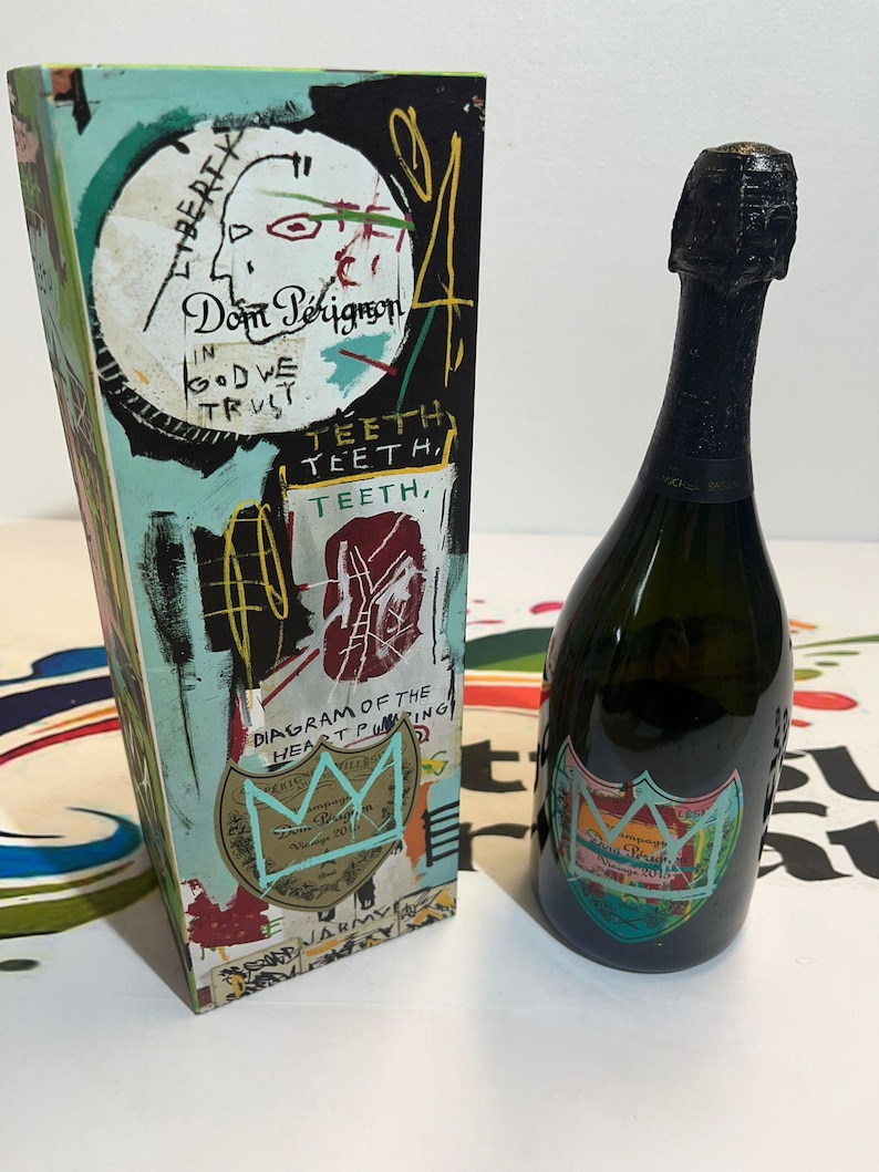 Pu&ograve; includere: Una bottiglia di champagne Dom P&eacute;rignon con una parte superiore in lamina nera e un'etichetta colorata. Accanto, una scatola abbinata con opere d'arte in stile graffiti, tra cui testo e un disegno a corona. La scatola riporta le parole "Dom P&eacute;rignon" e "In God We Trust".