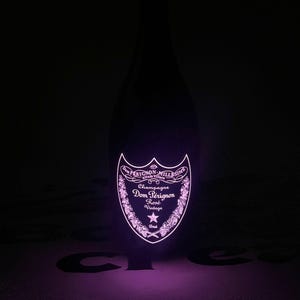 Może przedstawiać: Ciemna szklana butelka szampana Dom P&eacute;rignon Ros&eacute; Vintage z jarzącą się, podświetlaną etykietą w żywym r&oacute;żowym odcieniu. Etykieta zawiera napis Dom P&eacute;rignon, Champagne, Ros&eacute;, Vintage oraz Brut wraz z ozdobnymi motywami kwiatowymi.