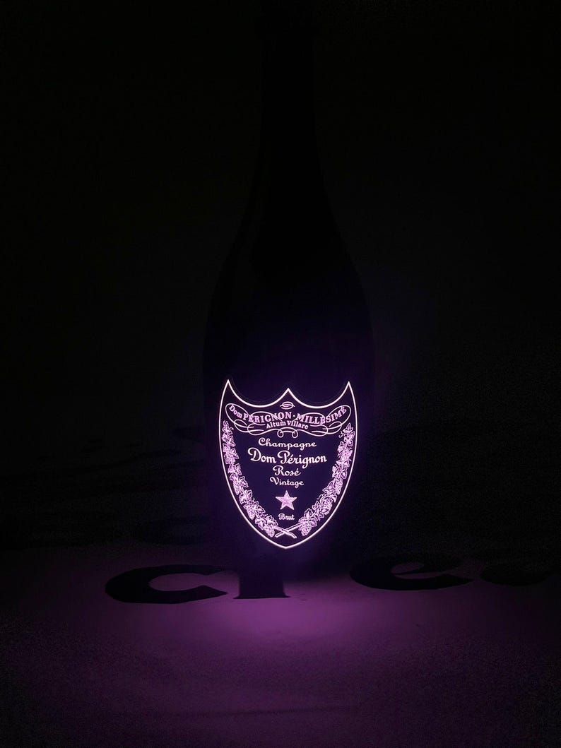 Może przedstawiać: Ciemna szklana butelka szampana Dom P&eacute;rignon Ros&eacute; Vintage z jarzącą się, podświetlaną etykietą w żywym r&oacute;żowym odcieniu. Etykieta zawiera napis Dom P&eacute;rignon, Champagne, Ros&eacute;, Vintage oraz Brut wraz z ozdobnymi motywami kwiatowymi.