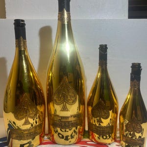 Puede incluir: Cuatro botellas de champán doradas de diferentes tamaños, cada una con un prominente emblema de as de picas en relieve en la parte frontal. Las botellas tienen cuellos envueltos en papel negro y etiquetas con el texto Armand de Brignac Brut Champagne.