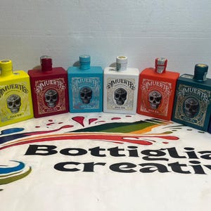 Può includere: Una collezione di bottiglie quadrate colorate, ognuna con un design a teschio e la parola "MUERTE". I colori includono nero, giallo, rosso e blu. Il testo "Bottiglia creativa" è visibile in primo piano.