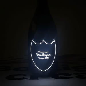 Może przedstawiać: Ciemna szklana butelka szampana Dom P&eacute;rignon Vintage 2015. Butelka posiada świecącą, podświetlaną etykietę w kształcie tarczy z przodu, na kt&oacute;rej wyraźnie widać napis Champagne Dom P&eacute;rignon Vintage 2015 w jasnym, białym świetle.