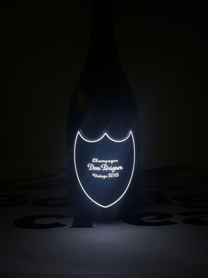Może przedstawiać: Ciemna szklana butelka szampana Dom P&eacute;rignon Vintage 2015. Butelka posiada świecącą, podświetlaną etykietę w kształcie tarczy z przodu, na kt&oacute;rej wyraźnie widać napis Champagne Dom P&eacute;rignon Vintage 2015 w jasnym, białym świetle.