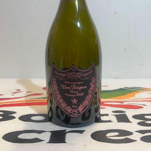 Może przedstawiać: Ciemnozielona szklana butelka szampana Dom P&eacute;rignon Ros&eacute; Vintage 2009. Etykieta ma kształt czarnej tarczy z r&oacute;żowymi kwiatowymi detalami, tekst Dom P&eacute;rignon Mill&eacute;sime, Champagne, Ros&eacute;, Vintage 2009, Brut, 750ml oraz 12,5% obj.