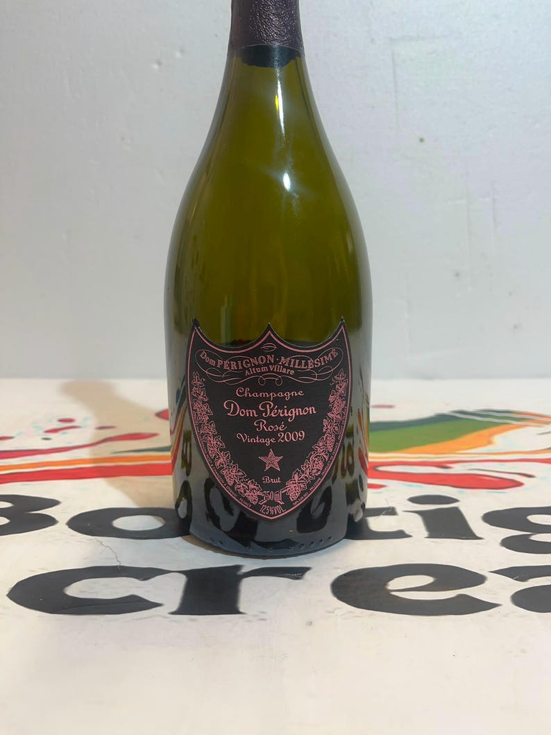 Może przedstawiać: Ciemnozielona szklana butelka szampana Dom P&eacute;rignon Ros&eacute; Vintage 2009. Etykieta ma kształt czarnej tarczy z r&oacute;żowymi kwiatowymi detalami, tekst Dom P&eacute;rignon Mill&eacute;sime, Champagne, Ros&eacute;, Vintage 2009, Brut, 750ml oraz 12,5% obj.