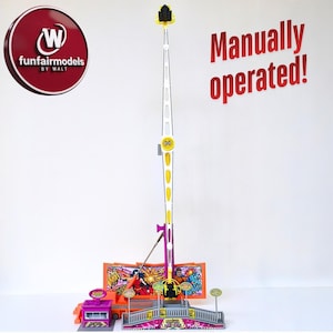 May include: A miniature fairground ride model featuring a tall, white and yellow tower. The model includes a colourful base with a ticket booth, bridge, and signs. The text "Manually operated!" is visible.