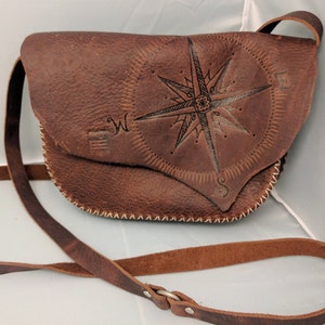 Puede incluir: Bolso bandolera de cuero marrón con diseño de rosa de los vientos. El bolso tiene un cierre de solapa y una correa larga y ajustable. La rosa de los vientos está grabada en el cuero. El bolso está cosido con hilo blanco.
