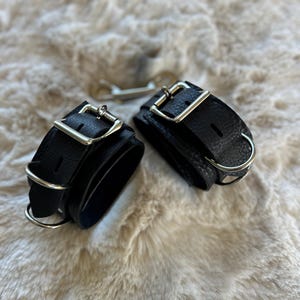 May include: Black leather wrist restraints with silver-colored buckles and rings. The restraints are lying on a soft, textured surface, and a silver-colored key is visible in the background.