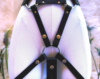 Puede incluir: Un arnés de cuero negro con una serie de correas interconectadas, anillos de metal plateado y remaches circulares. El diseño incluye una correa de cintura horizontal, dos correas diagonales que se unen en un anillo central y dos correas verticales para las piernas.