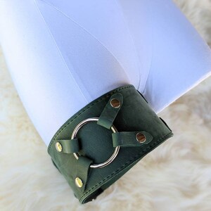 May include: A dark green leather cuff with a gold metal ring and gold metal rivets. The cuff is on a white fur surface.