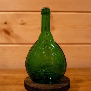 May include: A green glass bottle with a cork stopper, featuring an embossed design of a building. The bottle is displayed on a small, round wooden stand, set against a wooden background. The bottle's shape is bulbous.