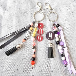 Op de afbeelding: Twee sleutelhangers met zilveren sluitingen, elk met een zwarte kwast, kralenaccenten en linten. Eén sleutelhanger heeft de letters "BTS" en de andere een cirkelvormig logo. De sleutelhangers hebben rode, roze en paarse accenten.
