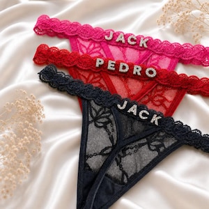 May include: Three thong panties in pink, red, and black lace with scalloped waistbands. Each pair features silver-toned rhinestone letters attached to the front waistband, spelling out the names JACK, PEDRO, and JACK respectively.