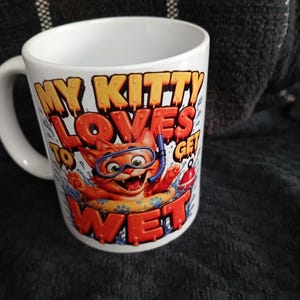 May include: A white ceramic mug featuring a colourful graphic of an orange cat wearing blue snorkelling goggles and floating in a yellow inner tube. The text reads MY KITTY LOVES TO GET WET in bold orange and red letters with a dripping effect.