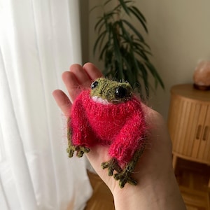 Könnte beinhalten: Eine kleine, flauschige, olivgrüne gehäkelte Froschfigur, die einen leuchtend pinken, langärmeligen, fusseligen Pullover trägt. Der Frosch hat große, runde, schwarze Kunststoffaugen und detaillierte Füße mit mehreren Zehen sowie eine sichtbare weiße Naht entlang des Mundes.