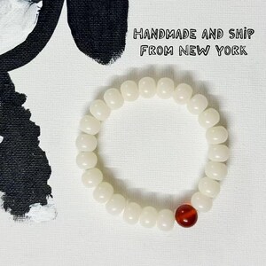 Può includere: Un braccialetto di perline bianche con una singola perla rossa. Il braccialetto è posto su uno sfondo bianco con un'illustrazione di cane dipinta in bianco e nero. Il testo nell'immagine recita "HANDMADE AND SHIP FROM NEW YORK."
