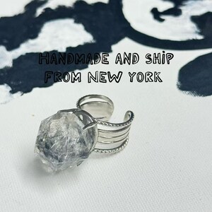 Pu&ograve; includere: Un anello regolabile color argento con un grande cristallo trasparente di forma irregolare. La fascia dell'anello ha un design a strati con un bordo strutturato. Lo sfondo include il testo "HANDMADE AND SHIP FROM NEW YORK" e un design astratto in bianco e nero.