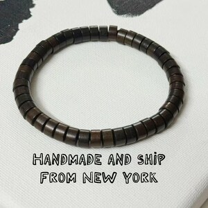 Può includere: Braccialetto con perline di legno marrone scuro. Il braccialetto è composto da piccole perline rettangolari infilate insieme. Lo sfondo è una tela bianca con un dipinto astratto scuro. Il testo "HANDMADE AND SHIP FROM NEW YORK" è in basso.