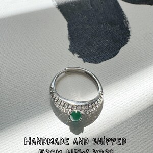 Può includere: Un anello d'argento con una piccola gemma verde rotonda al centro. La fascia è ornata da piccole pietre trasparenti. L'anello è posto su uno sfondo bianco con un motivo grigio scuro.