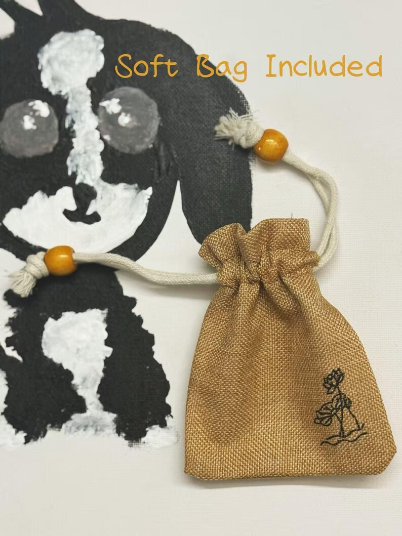 Pu&ograve; includere: Un piccolo sacchetto con coulisse beige con ricamo floreale, accompagnato da un cordoncino color crema e perline di legno. Le parole "Soft Bag Included" sono scritte in arancione. Un'illustrazione di cane in bianco e nero &egrave; sullo sfondo.