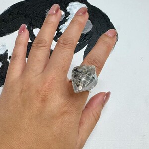 Pu&ograve; includere: Un anello d'argento con un grande cristallo trasparente di forma irregolare. Il cristallo presenta inclusioni scure ed &egrave; montato su una sottile fascia d'argento. L'anello &egrave; esposto su uno sfondo bianco con un dipinto astratto in bianco e nero.