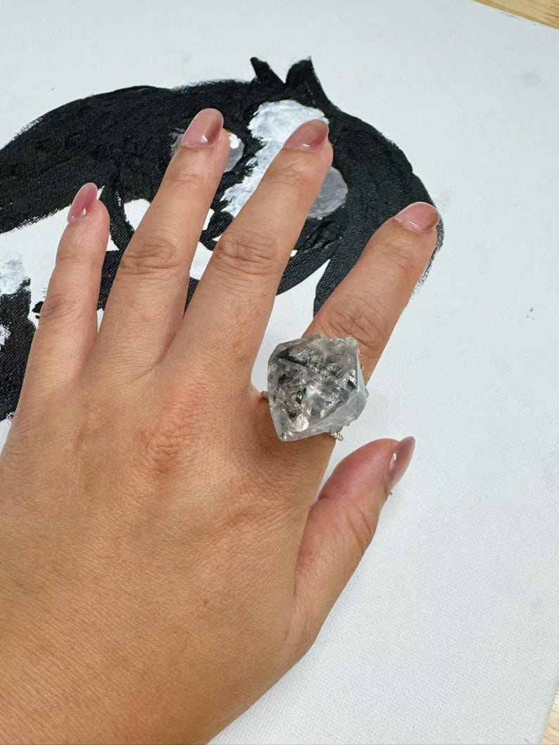 Pu&ograve; includere: Un anello d'argento con un grande cristallo trasparente di forma irregolare. Il cristallo presenta inclusioni scure ed &egrave; montato su una sottile fascia d'argento. L'anello &egrave; esposto su uno sfondo bianco con un dipinto astratto in bianco e nero.