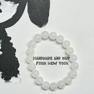Può includere: Un braccialetto di perline bianche composto da perline rotonde e opache. Il braccialetto è esposto su una superficie bianca con un disegno astratto in bianco e nero sullo sfondo. Il testo sull'immagine recita "HANDMADE AND SHIP FROM NEW YORK."