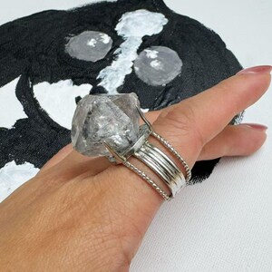 Pu&ograve; includere: Un grande anello di cristallo trasparente con una fascia argentata. L'anello presenta un cristallo grezzo e non tagliato incastonato in un design a pi&ugrave; bande. Il cristallo &egrave; tenuto in posizione da rebbi. L'anello &egrave; su una mano, con uno sfondo dipinto in bianco e nero.