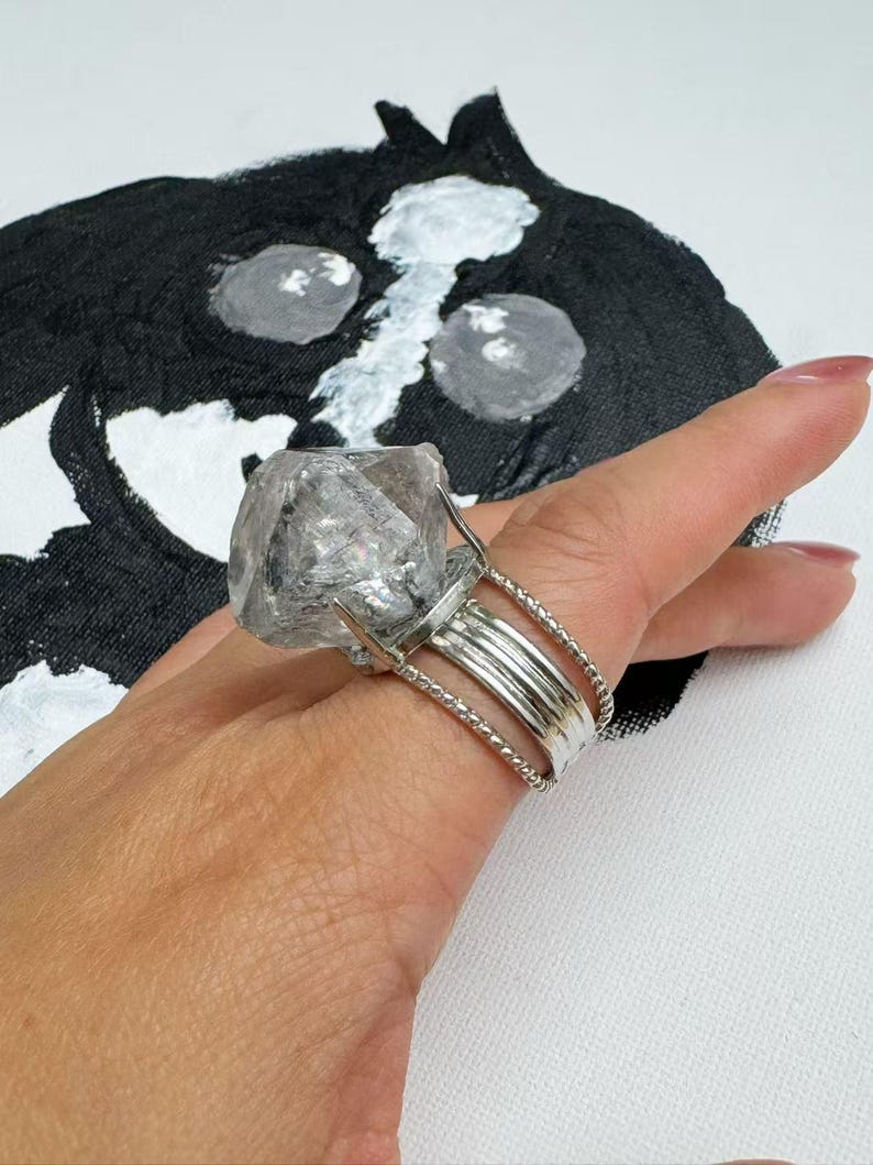 Pu&ograve; includere: Un grande anello di cristallo trasparente con una fascia argentata. L'anello presenta un cristallo grezzo e non tagliato incastonato in un design a pi&ugrave; bande. Il cristallo &egrave; tenuto in posizione da rebbi. L'anello &egrave; su una mano, con uno sfondo dipinto in bianco e nero.