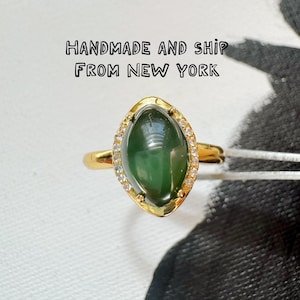 Può includere: Un anello d'oro con una grande pietra preziosa verde ovale, circondata da piccole pietre trasparenti. L'anello ha una fascia in oro lucido. Il testo "HANDMADE AND SHIP FROM NEW YORK" è visibile. Un gioiello.