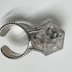 Pu&ograve; includere: Anello regolabile color argento con un grande cristallo di quarzo trasparente sfaccettato con inclusioni scure. La fascia dell'anello ha un bordo decorativo bordato. Il quarzo &egrave; tagliato a forma geometrica.