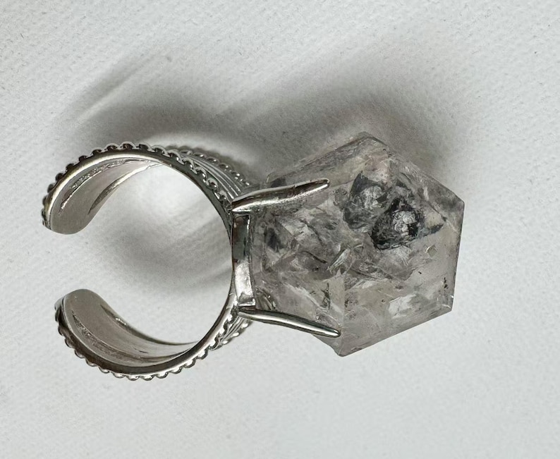 Pu&ograve; includere: Anello regolabile color argento con un grande cristallo di quarzo trasparente sfaccettato con inclusioni scure. La fascia dell'anello ha un bordo decorativo bordato. Il quarzo &egrave; tagliato a forma geometrica.