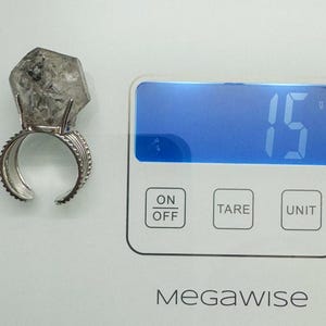 Pu&ograve; includere: Un anello d'argento con una grande pietra di cristallo trasparente di forma irregolare. L'anello &egrave; su una bilancia digitale, che indica 15 grammi. La bilancia ha un pulsante on/off, un pulsante tara e un pulsante unit&agrave;. Il marchio "MegaWise" &egrave; stampato sulla bilancia.