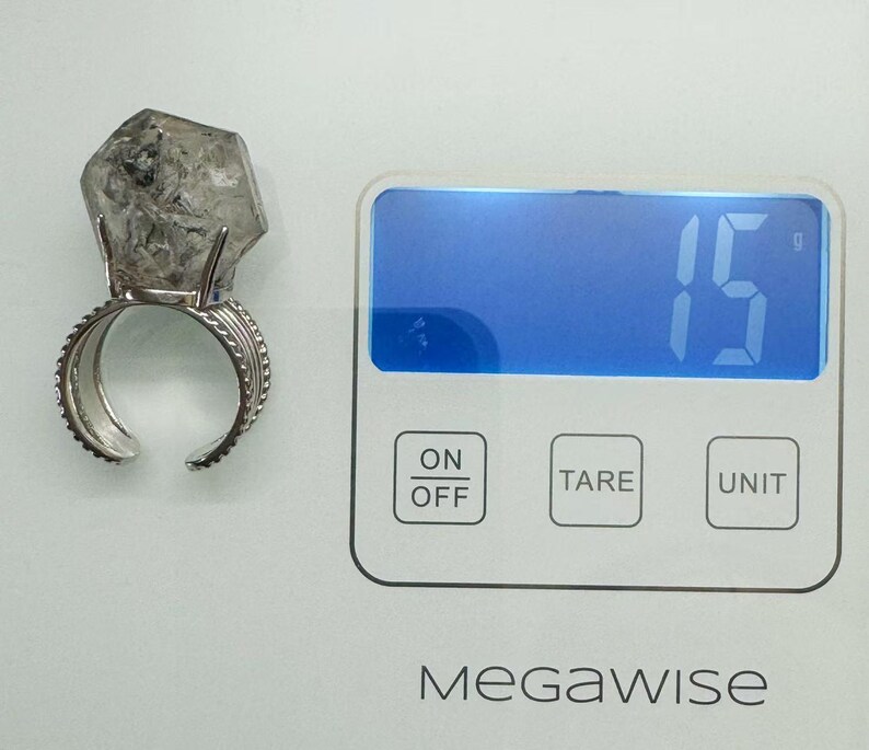 Pu&ograve; includere: Un anello d'argento con una grande pietra di cristallo trasparente di forma irregolare. L'anello &egrave; su una bilancia digitale, che indica 15 grammi. La bilancia ha un pulsante on/off, un pulsante tara e un pulsante unit&agrave;. Il marchio "MegaWise" &egrave; stampato sulla bilancia.