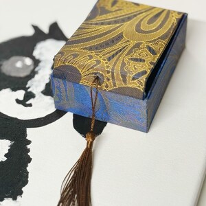 Pu&ograve; includere: Una confezione regalo rettangolare con un motivo paisley blu e oro. La scatola ha un nappa marrone attaccata al coperchio. La scatola &egrave; su una superficie bianca con un'illustrazione in bianco e nero sullo sfondo.