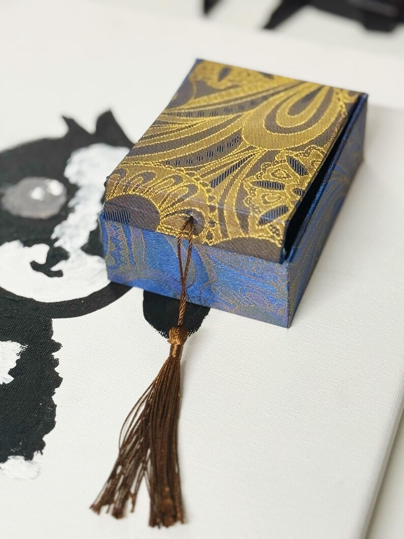Pu&ograve; includere: Una confezione regalo rettangolare con un motivo paisley blu e oro. La scatola ha un nappa marrone attaccata al coperchio. La scatola &egrave; su una superficie bianca con un'illustrazione in bianco e nero sullo sfondo.
