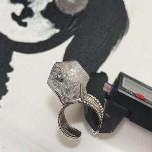 Pu&ograve; includere: Un anello argentato con una grande gemma esagonale trasparente. La fascia dell'anello ha un design strutturato. Un calibro digitale &egrave; parzialmente visibile, suggerendo le dimensioni dell'anello.