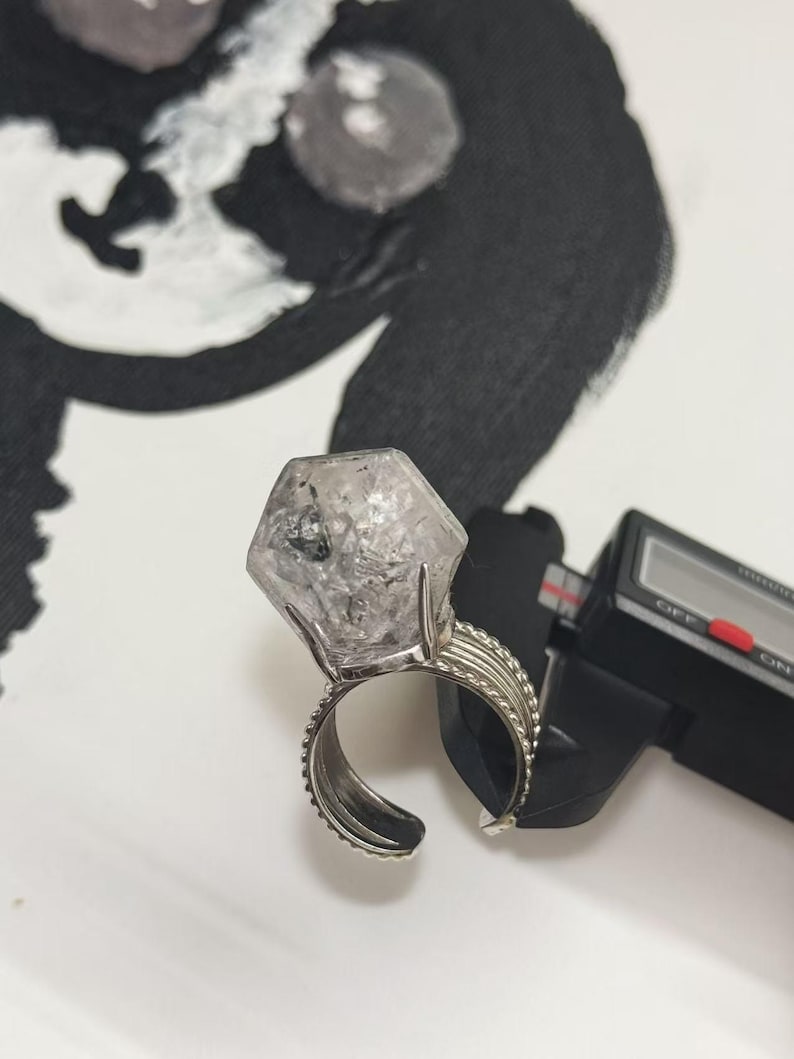 Pu&ograve; includere: Un anello argentato con una grande gemma esagonale trasparente. La fascia dell'anello ha un design strutturato. Un calibro digitale &egrave; parzialmente visibile, suggerendo le dimensioni dell'anello.
