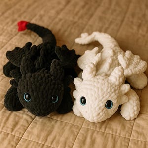 May include: Two crocheted dragon toys, one black and one white, are shown together. The black dragon has a red tail tip and blue eyes. The white dragon has blue eyes. Both are made of a soft, textured material.