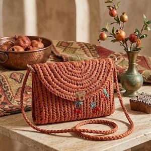 May include: A woven, rust-colored crossbody bag with a flap closure. The bag features a decorative cherry design with red and green accents. The bag has a braided strap and a gold-colored clasp.
