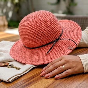 May include: A coral-colored crocheted sun hat with a rounded crown and a wide brim. A thin, dark brown braided cord is tied around the base of the crown, featuring a small brown leather-like tag on the side of the brim. The hat has a textured finish.