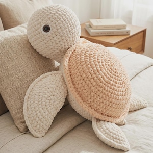 May include: A plush, crocheted turtle in cream and peach. The turtle has a round head with a single brown eye, a peach-colored shell, and cream-colored flippers. The turtle is lying on a bed with neutral-colored bedding.