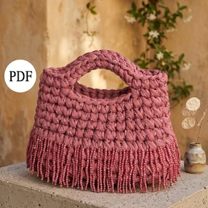 May include: A dusty rose-colored handbag with a braided handle and beaded fringe. The bag is made of a thick, textured material and has a rounded shape. The word "PDF" is in a white circle on the bag.