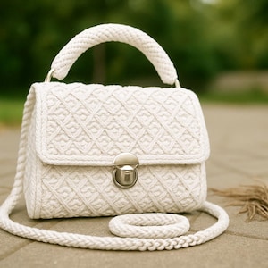 May include: Cream-colored handbag with a woven pattern, a top handle, and a long shoulder strap. The bag has a silver-colored clasp and a matching bracelet. A small bundle of tan-colored fibers rests beside the bag. The bag is made of a soft, woven material.